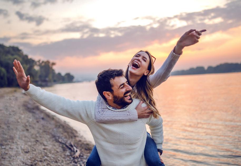 A man gives a woman a piggyback ride on a lakeshore at sunset. Both are smiling and laughing, with arms outstretched, enjoying the scenic view and the moment together.
