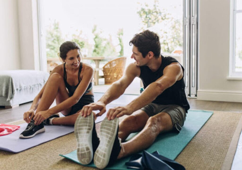 Two people in athletic wear sit on yoga mats indoors, stretching and smiling at each other in a bright room with large windows and outdoor views.