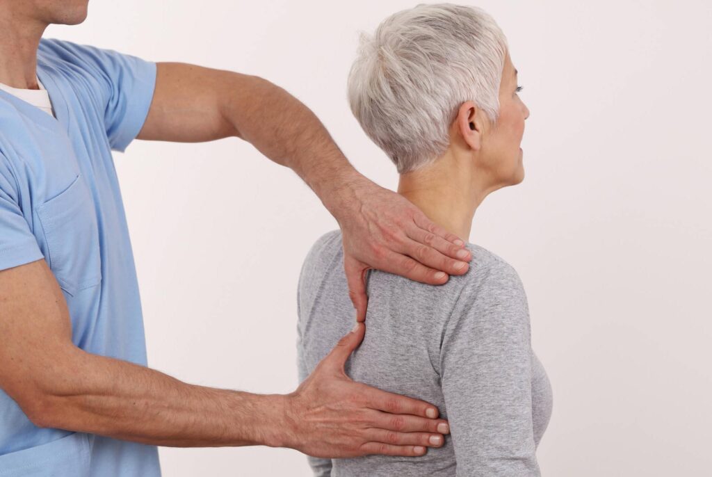 A healthcare professional gently examines the upper back and shoulders of an older woman with short gray hair, who is wearing a gray long-sleeve shirt, against a plain background.