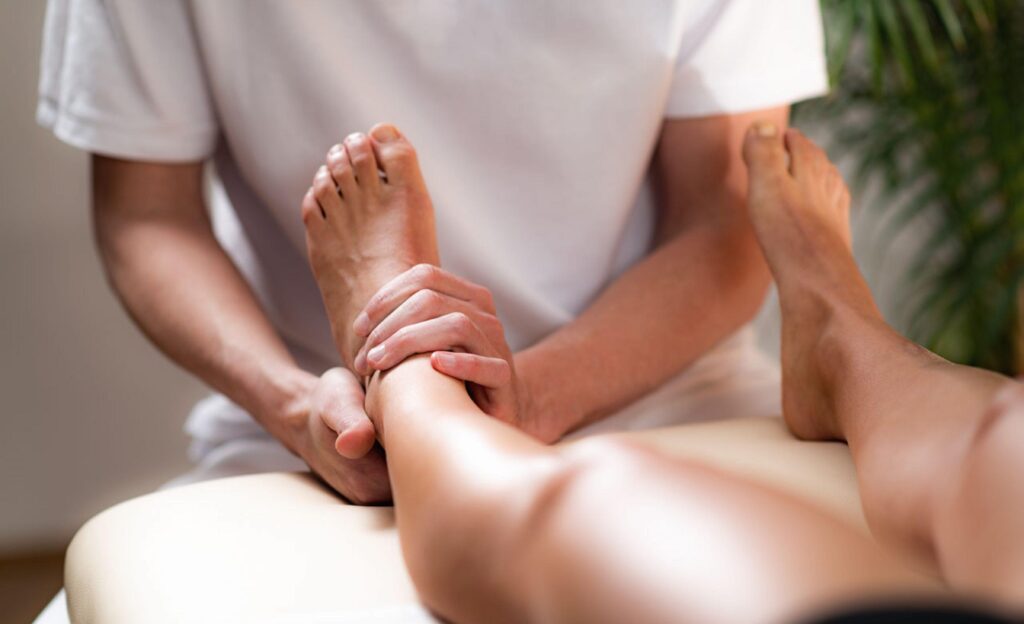 A person in a white shirt gives a foot and ankle massage to someone lying on a massage table, with a soft background and greenery visible.