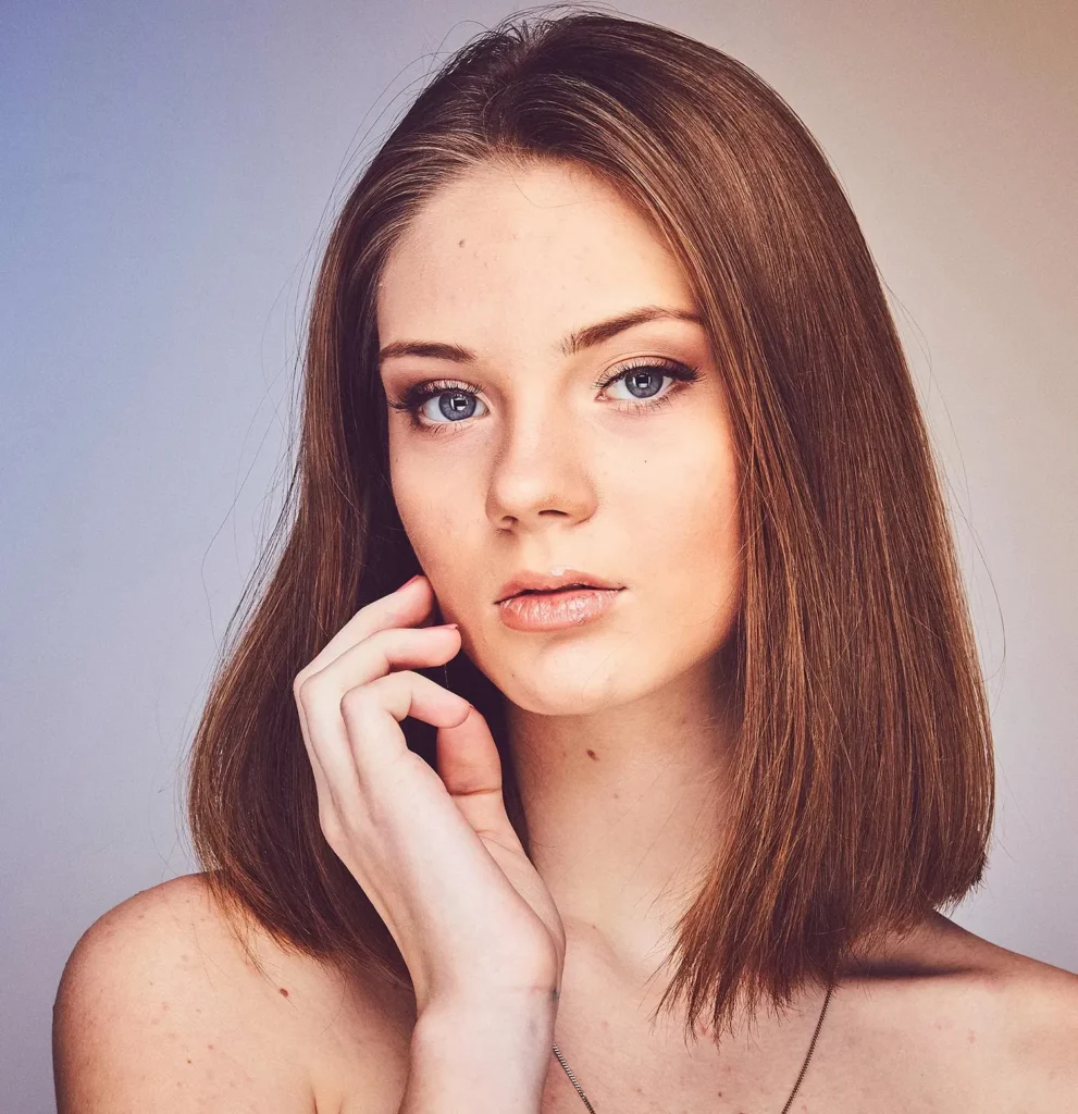 A young woman with straight, shoulder-length brown hair and blue eyes gazes at the camera, gently touching her face with one hand against a neutral background.
