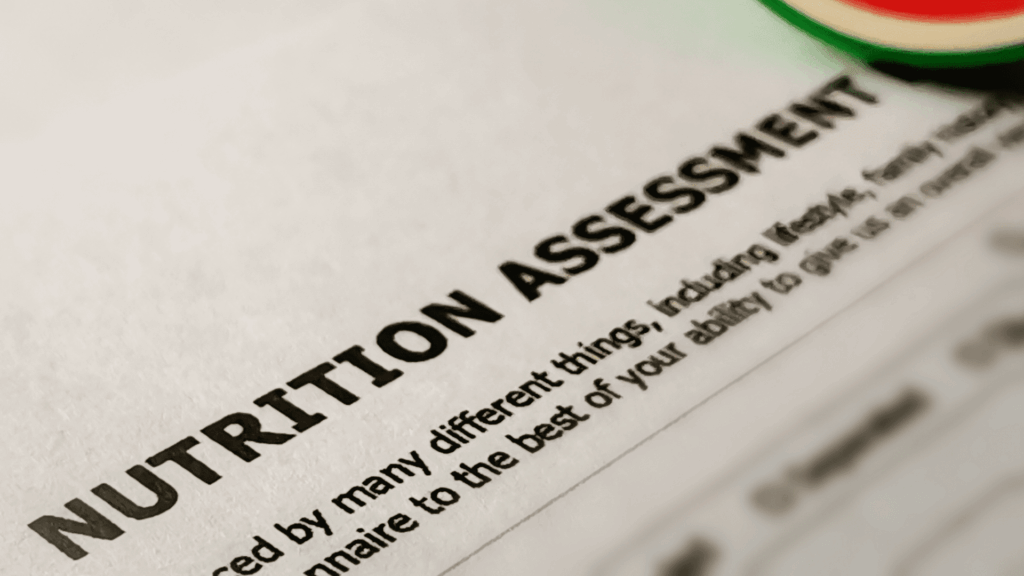 Close-up of a document titled NUTRITION ASSESSMENT in bold black letters, showing part of the printed text and a blurred section of a circular object in the top right corner.